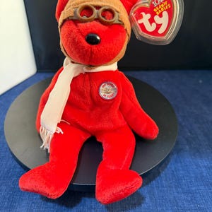 Puede incluir: Un oso de peluche rojo con una gorra de aviador marrón con gafas y una bufanda blanca. El oso tiene una nariz negra y un pin circular que dice "100 Years of Flight". Una etiqueta en forma de corazón dice "Ty Beanie Babies".