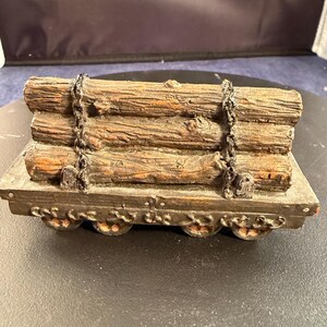 May include: A miniature wooden log cart with four wheels. The cart bed is a dark gray color, and it holds a stack of wooden logs. The logs are secured with a chain. The cart is a decorative item.