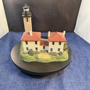 May include: A miniature lighthouse figurine featuring a tall stone-coloured tower with a black lantern room, connected to two cream-coloured buildings with red tiled roofs. The structure sits on a green base with a small yellow label marked 4734.