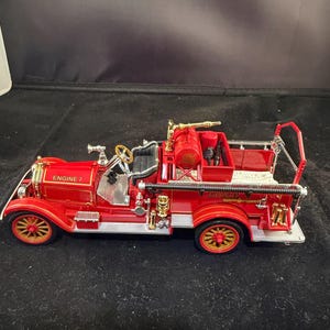 May include: A detailed red antique fire engine model with gold detailing. The model features the text "ENGINE 7" on the side and has gold-coloured wheels. The fire engine has a ladder and other equipment on the back.