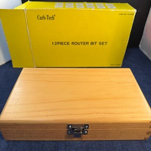 May include: A wooden box with a metal clasp and a yellow box with the text "Carb-Tech" and "12 PIECE ROUTER BIT SET". The boxes are on a blue surface.