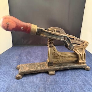 May include: Antique cast iron potato cutter with a wooden handle. The cutter is painted in a dark color and features a spring mechanism. The handle is a reddish-brown color. The base of the cutter has an ornate design. The text "ENTERPRISE NO. 9" is visible.