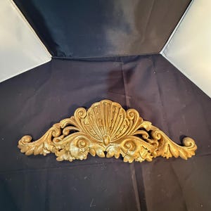 Puede incluir: Un frontón decorativo de pared en color dorado que presenta un motivo central de concha flanqueado por patrones simétricos de hojas de acanto en espiral. La pieza tiene un acabado metálico texturizado con detalles en relieve.