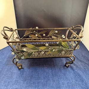 May include: A decorative metal tray with a rectangular shape, featuring ornate scrollwork and leaf accents. The tray has a bronze finish with green and clear bead embellishments. It rests on four scrolled feet and has handles.