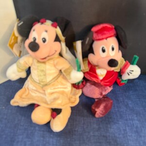 May include: Two plush Disney character dolls. One doll is dressed in a yellow dress with gold trim and a matching headpiece. The other doll is dressed in a red velvet outfit with a matching hat. Both dolls have black ears and white gloves.