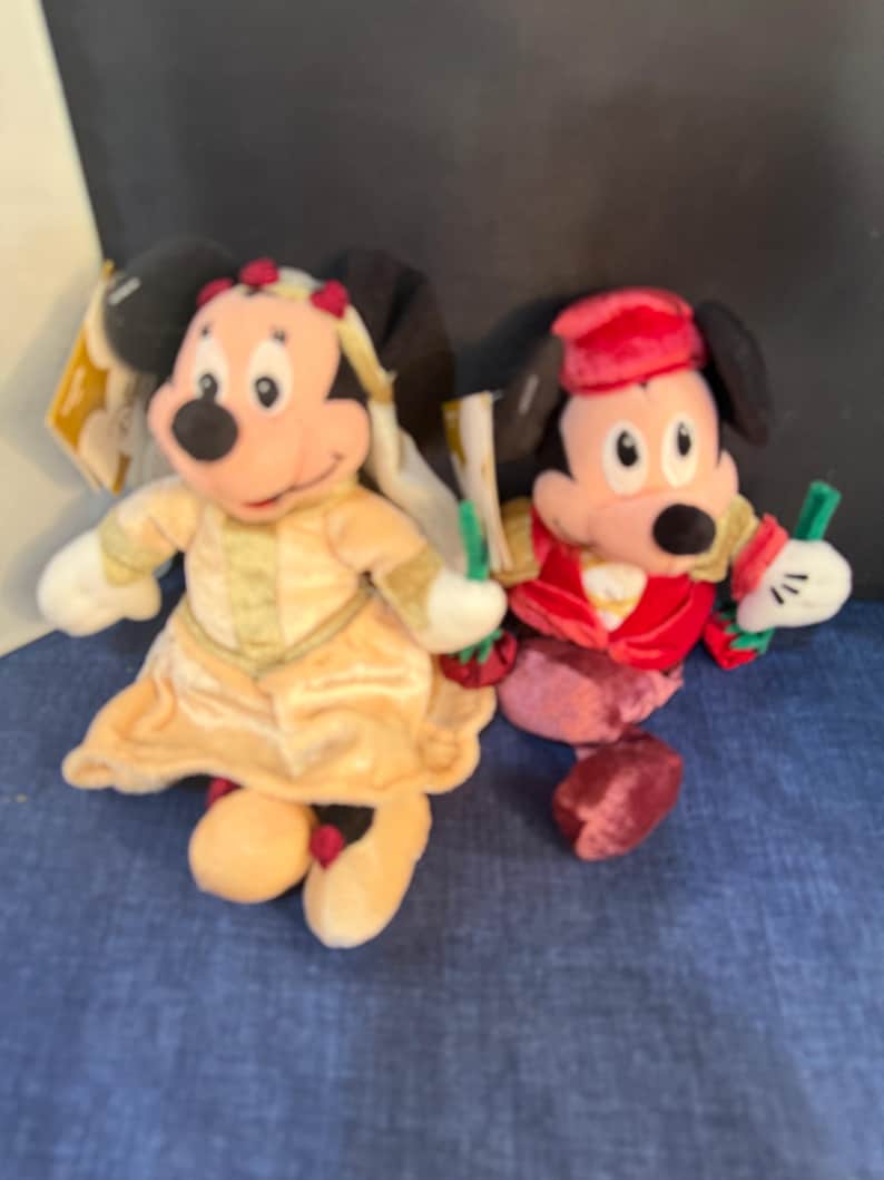 May include: Two plush Disney character dolls. One doll is dressed in a yellow dress with gold trim and a matching headpiece. The other doll is dressed in a red velvet outfit with a matching hat. Both dolls have black ears and white gloves.