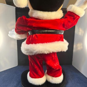 May include: A Mickey Mouse Christmas figurine, viewed from the back. Mickey is dressed in a red velvet Santa suit with white faux fur trim. He wears a Santa hat and has white gloves. The figurine stands on a black base.