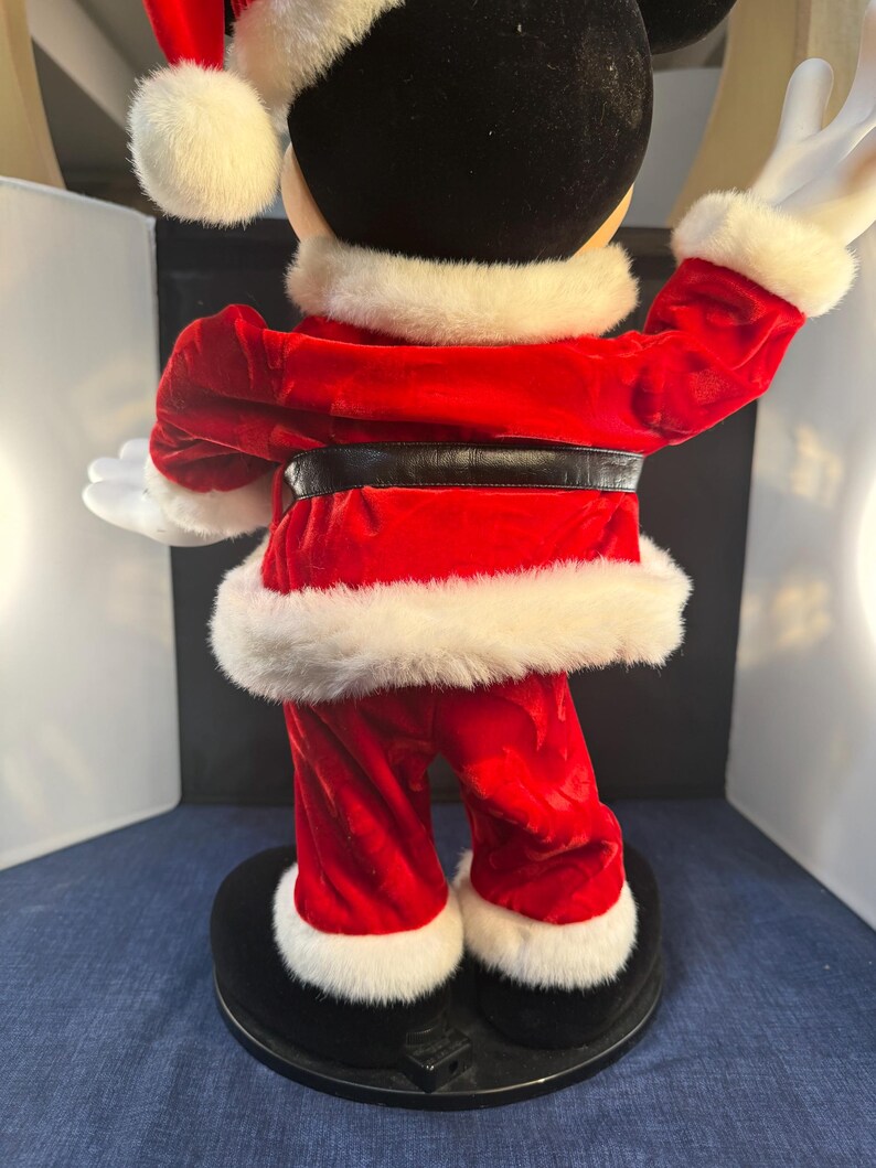 May include: A Mickey Mouse Christmas figurine, viewed from the back. Mickey is dressed in a red velvet Santa suit with white faux fur trim. He wears a Santa hat and has white gloves. The figurine stands on a black base.