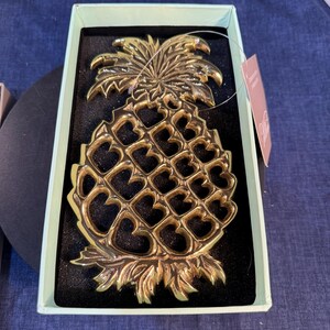 May include: A gold-toned metal pineapple-shaped trivet with a cut-out lattice design featuring heart-shaped openings. The trivet is nestled inside a light green rectangular box with a black foam insert and includes a small attached paper tag.