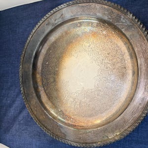 Peut inclure: Un plateau de service rond en métal argenté avec un motif floral en relief détaillé au centre. Le plateau a un bord décoratif torsadé en forme de corde. La surface montre des signes d'usure, ajoutant à son attrait vintage.