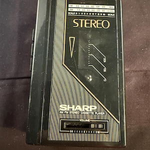 Könnte beinhalten: Ein schwarzer Sharp AM-FM Stereo-Kassettenrekorder mit goldfarbenen Akzenten und Schriftzügen. Das Gerät verfügt über eine Radiofrequenzanzeige für FM- und AM-Bänder, einen Lautstärkeregler von 0 bis 10 und ein Sichtfenster für die Kassette.