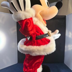 May include: A plush Mickey Mouse figure dressed in a Santa Claus outfit. The red velvet suit is trimmed with white faux fur. The figure has black shoes, a black belt with a gold buckle, and white gloves. The figure is in a celebratory pose.