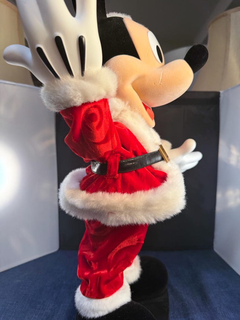 May include: A plush Mickey Mouse figure dressed in a Santa Claus outfit. The red velvet suit is trimmed with white faux fur. The figure has black shoes, a black belt with a gold buckle, and white gloves. The figure is in a celebratory pose.