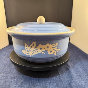 Pode incluir: Uma travessa de cerâmica azul clara com tampa e botão branco. A travessa tem um desenho floral branco e uma borda creme. Assenta sobre uma base circular preta. O fundo é uma superfície azul.