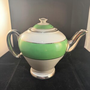 Puede incluir: Una tetera vintage con un diseño blanco, verde y plateado. La tetera tiene un cuerpo redondeado con una banda verde, una parte superior blanca y una boquilla y asa plateadas. La tapa es verde y blanca con un asa plateada.