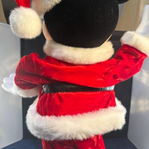 May include: A Mickey Mouse figure dressed in a Santa Claus outfit, viewed from the back. The red velvet suit has white faux fur trim, a black belt, and a matching Santa hat. The figure's black head and white gloves are visible.