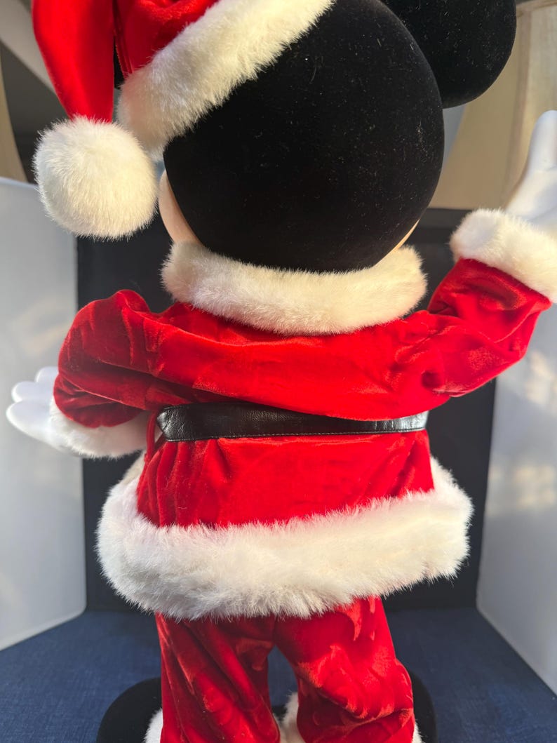 May include: A Mickey Mouse figure dressed in a Santa Claus outfit, viewed from the back. The red velvet suit has white faux fur trim, a black belt, and a matching Santa hat. The figure's black head and white gloves are visible.