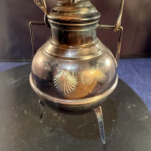 May include: A tarnished silver-toned metal vessel with a rounded body, a lid, and a swing handle. The container features three thin legs and a decorative embossed seashell motif on the front. The surface shows signs of oxidation and dark patina.