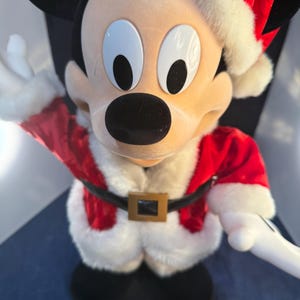 May include: A Mickey Mouse figure dressed in a Santa Claus outfit. The figure has a red jacket with white trim, a black belt with a gold buckle, and a Santa hat. The figure is standing on a black base and has a cheerful expression.