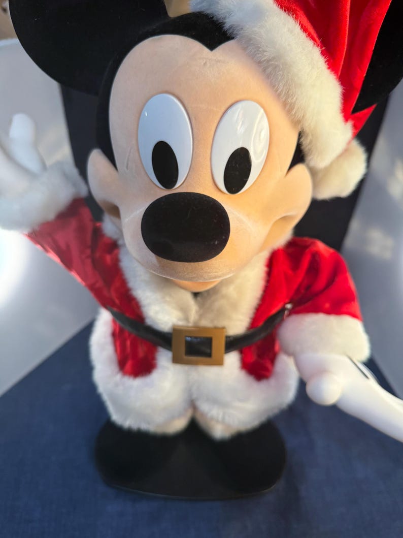 May include: A Mickey Mouse figure dressed in a Santa Claus outfit. The figure has a red jacket with white trim, a black belt with a gold buckle, and a Santa hat. The figure is standing on a black base and has a cheerful expression.