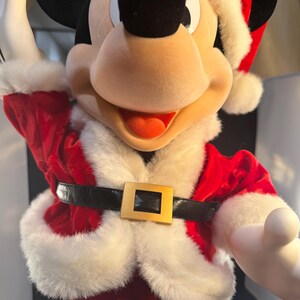 May include: Mickey Mouse dressed in a Santa Claus outfit. The red velvet suit has white faux fur trim and a black belt with a gold buckle. Mickey's black ears and face contrast with his white gloves and Santa hat.