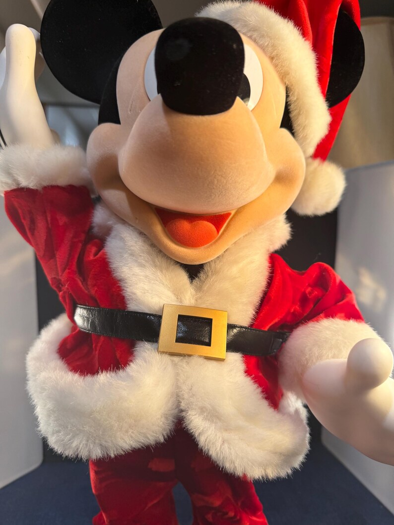 May include: Mickey Mouse dressed in a Santa Claus outfit. The red velvet suit has white faux fur trim and a black belt with a gold buckle. Mickey's black ears and face contrast with his white gloves and Santa hat.