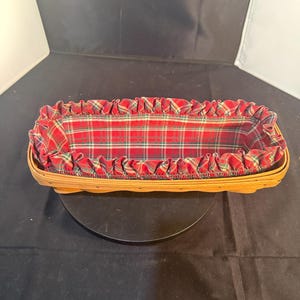 May include: A rectangular woven wooden basket featuring a light brown natural finish. The interior is lined with a red, green, and white plaid fabric that extends over the top rim in a gathered, ruffled edge, creating a decorative border.