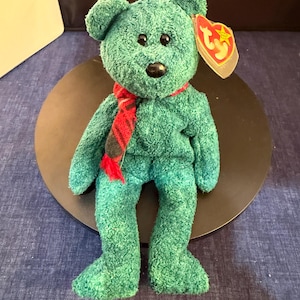 Puede incluir: Un oso de peluche verde azulado con nariz y ojos negros, que lleva una bufanda a cuadros roja y negra. El oso tiene una etiqueta Ty adjunta a su oreja.