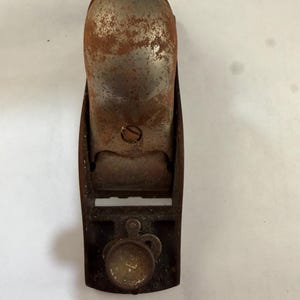 Puede incluir: Un cepillo de mano antiguo, que muestra signos de edad con óxido y desgaste. La herramienta está hecha de metal y presenta una perilla de ajuste de la cuchilla. El cuerpo del cepillo es de color oscuro y desgastado, con una parte superior curva y una base plana. Una herramienta de carpintería vintage.