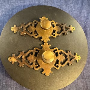 May include: Two brass drawer pulls featuring ornate, symmetrical scrollwork backplates with a central circular knob. Each piece has a weathered, metallic finish with intricate cut-out details and small screw holes at the pointed ends for mounting.