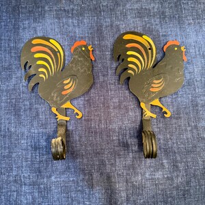 May include: Two black metal rooster-shaped wall hooks. The roosters have orange, yellow, and black tail feathers and red combs. The hooks are attached to a dark metal base. The hooks are on a blue background.
