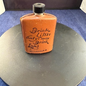 May include: A vintage brown leather flask with a black cap. The flask has the text "Drink Little that ye may Drink Long" in black script. The flask is rectangular and appears to be made of leather.