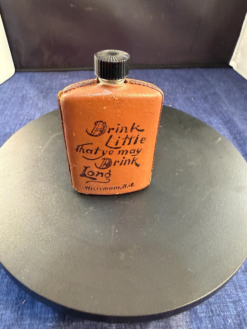 May include: A vintage brown leather flask with a black cap. The flask has the text "Drink Little that ye may Drink Long" in black script. The flask is rectangular and appears to be made of leather.