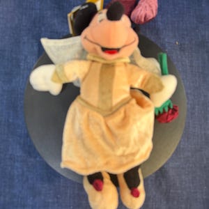 May include: A plush Minnie Mouse doll wearing a light orange dress with a matching collar and shoes. The doll has white gloves, a black head with a red bow, and is holding a green and red item. The doll is on a dark gray surface.
