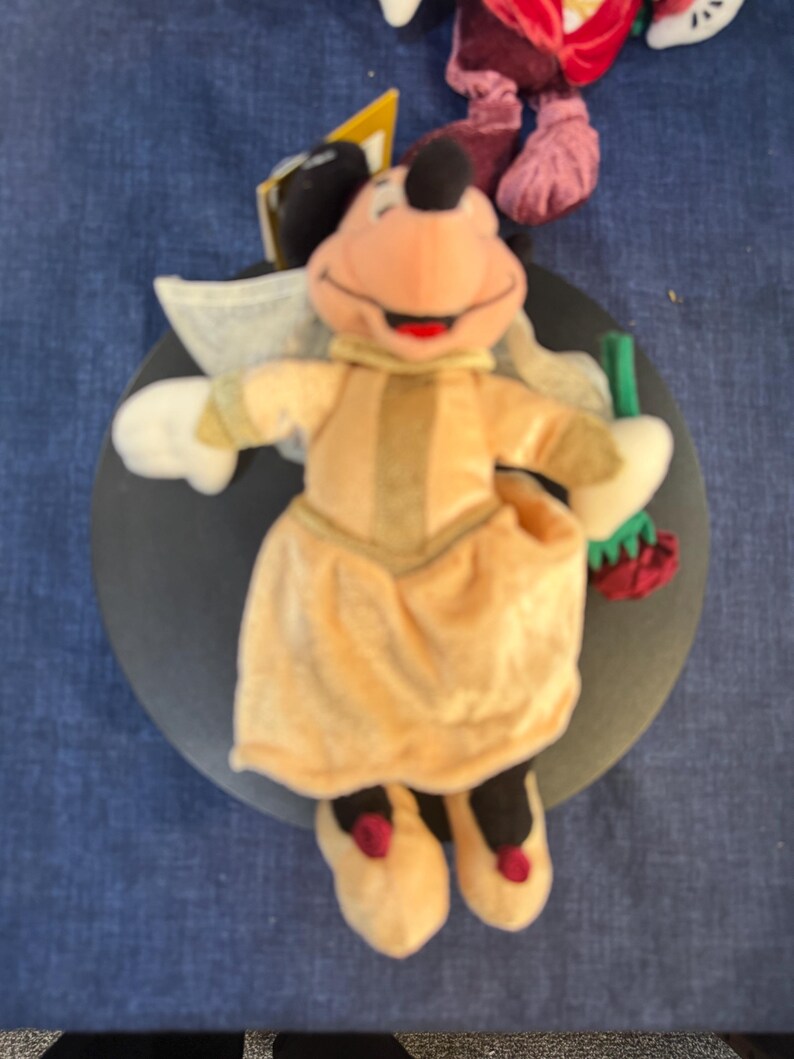 May include: A plush Minnie Mouse doll wearing a light orange dress with a matching collar and shoes. The doll has white gloves, a black head with a red bow, and is holding a green and red item. The doll is on a dark gray surface.