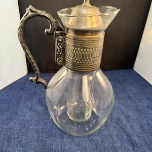 Puede incluir: Una jarra de vidrio transparente y plateada con asa y tapa decorativas. La jarra tiene una base bulbosa y una banda plateada alrededor del centro con un diseño ornamentado. La tapa está rematada con un adorno plateado.