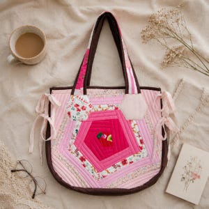 May include: A patchwork tote bag in various shades of pink and brown, with a geometric pattern. The bag has brown trim, handles, and decorative bows. Floral fabric details and a small hexagon patch are visible. A cup of coffee, glasses, and a book are in the background.