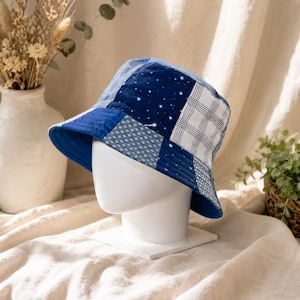May include: A patchwork bucket hat in shades of blue, white, and denim. The hat features a variety of patterned squares, including a starry night design. The hat is displayed on a white mannequin head.