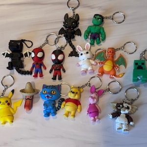 May include: A collection of thirteen character keychains featuring various figures including a black dragon, green superhero, red and blue spider-themed hero, white rabbit, orange dragon, green block creature, yellow electric mouse, and a brown bear.