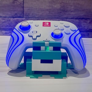 May include: A white Nintendo Switch Pro controller with blue wave patterns and glowing blue joysticks resting on a teal and white block-style stand. The controller features the red and white Nintendo Switch logo in the center.