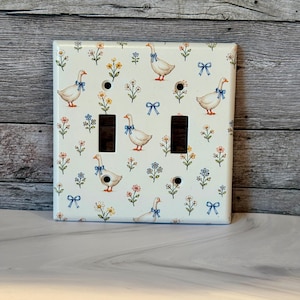 May include: A cream-colored double light switch wall plate featuring a repeating pattern of white geese wearing blue bows, interspersed with small yellow, pink, and blue flowers and blue ribbon bows. The plate has two rectangular cutouts.