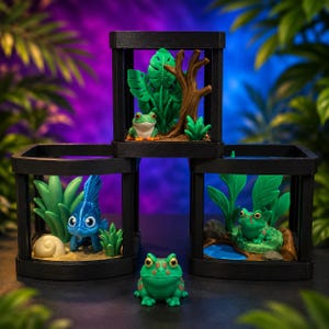 May include: Three black square display frames containing miniature dioramas. One frame holds a frog with green leaves and a tree branch. Another holds a blue fish with a shell. The third holds a green frog with a small blue pond. A loose green frog sits in front.