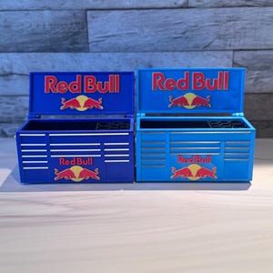 May include: Two miniature tool chests in dark blue and light blue, each featuring the Red Bull logo and text on the lid and front. The chests have open lids, multiple drawer fronts with silver accents, and a black interior compartment with circular slots.