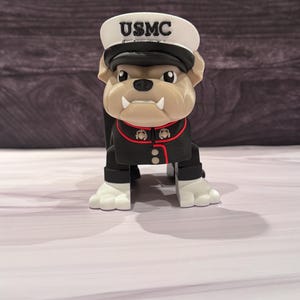 May include: A figurine of a bulldog wearing a white cap with the text USMC in black letters. The dog is dressed in a black military-style jacket with red trim, gold-colored buttons, and white paws. The figure has a tan face with two visible white fangs.