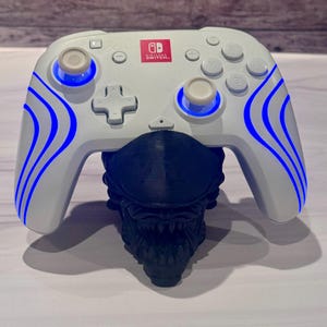 May include: A white Nintendo Switch wireless controller with blue LED light accents on the grips, resting on a black 3D-printed stand shaped like a creature head with visible teeth and a smooth, elongated cranium structure.