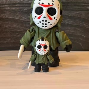 May include: Two figures of a character wearing a white hockey mask with red triangular markings, a green hooded jacket, and black pants. The larger figure holds a white blade in its right hand, while the smaller figure stands directly in front.