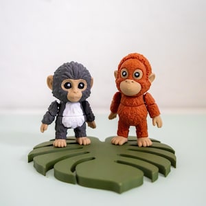 May include: Two small monkey figurines, one gray and one orange, stand on a green leaf-shaped base. The gray monkey has a white chest, while the orange monkey is a solid color. Both have large eyes and are in a playful pose.