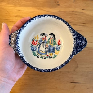 May include: A white ceramic bowl with a blue speckled rim and handles. The bowl features a colourful illustration of two figures in traditional clothing. The bowl is held in a person's hand.
