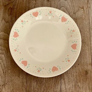 May include: A white ceramic plate with a decorative border featuring pink hearts and small floral accents. The plate is set against a wooden surface, showcasing its design.