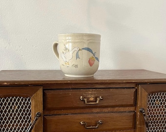 May include: A beige ceramic mug with a handle, featuring a goose holding a blue ribbon and a strawberry. The mug is set on a wooden cabinet with two drawers and a wire mesh door.